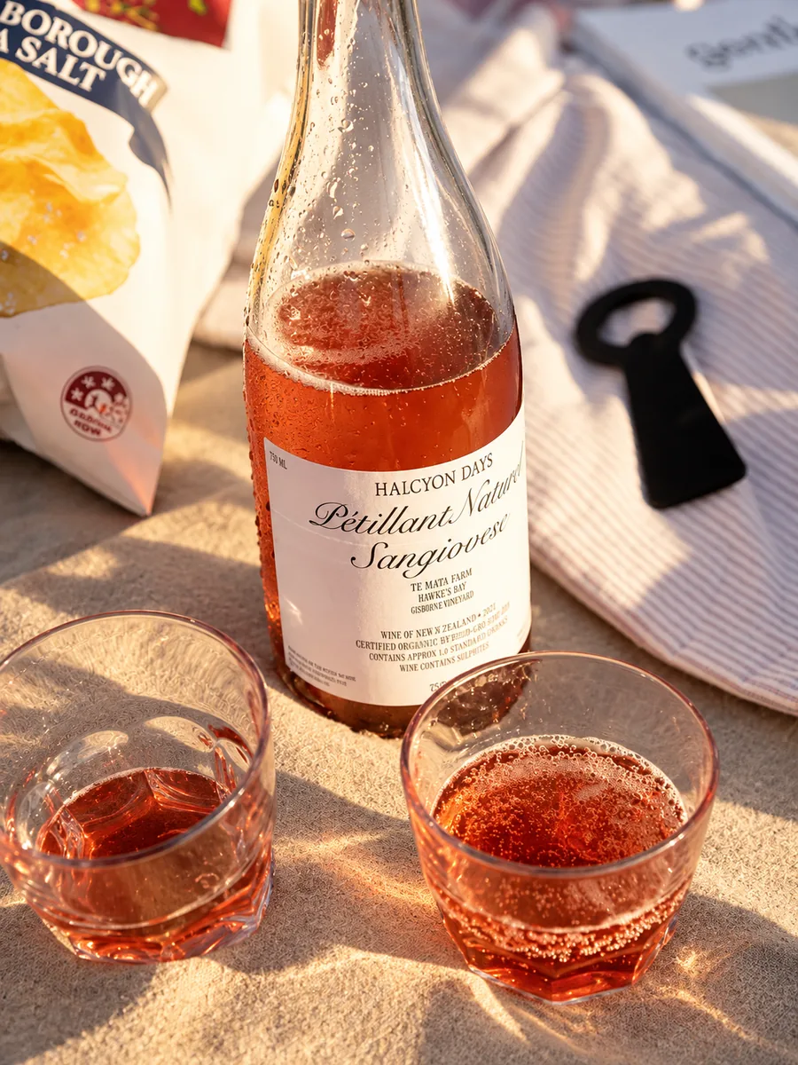 A bottle of pét-nat and glasses on a sunny table